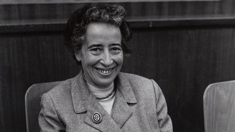 The picture shows a black-and-white photograph of Hannah Arendt. She is sitting on a chair and smiling warmly at the camera.