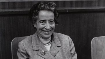 Das Bild zeigt eine Schwarz-Weiß-Aufnahme von Hannah Arendt. Sie sitzt auf einem Stuhl und lächelt herzlich in die Kamera.