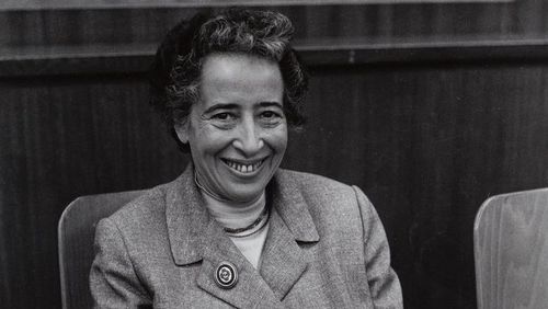 Das Bild zeigt eine Schwarz-Weiß-Aufnahme von Hannah Arendt. Sie sitzt auf einem Stuhl und lächelt herzlich in die Kamera.
