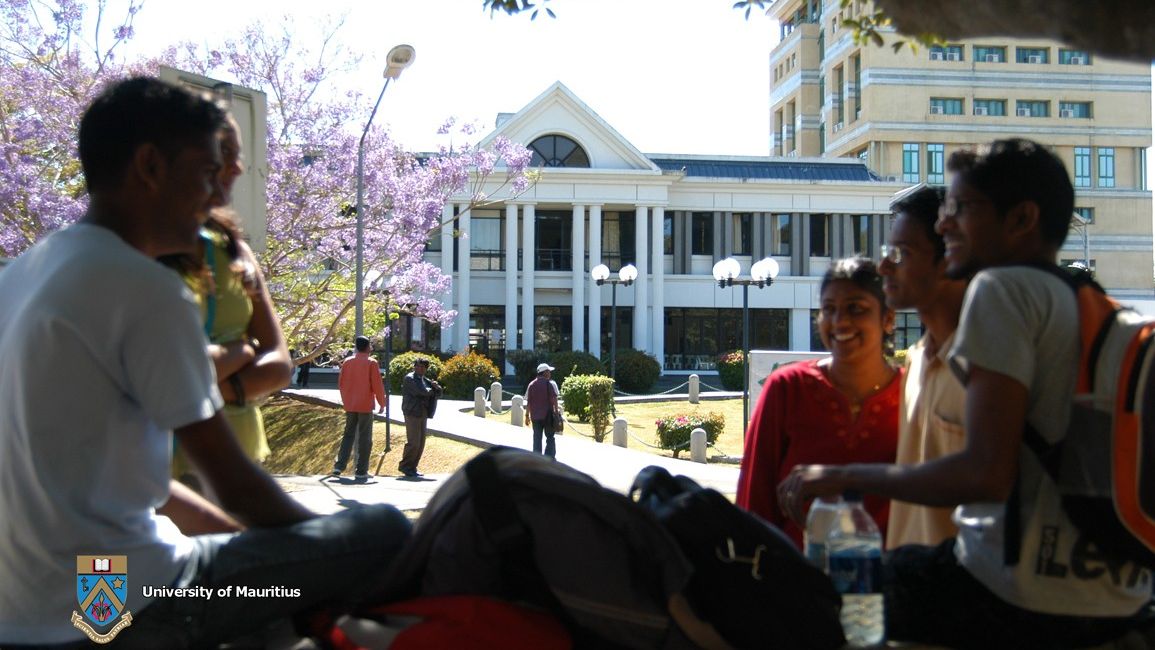 Students on a bench on the campus of the University of Mauritius. In the background, buildings with white facades & purple-coloured flowers.
