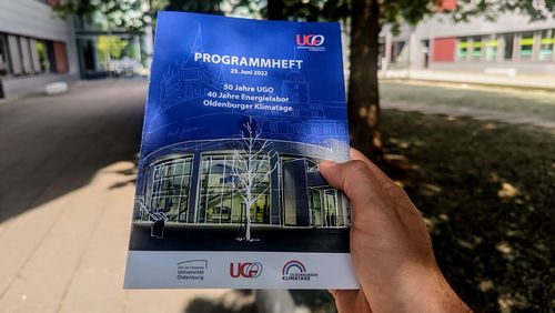 Das Programmheft wird von einer Hand gehalten, im Hintergrund Universitätsgebäude.