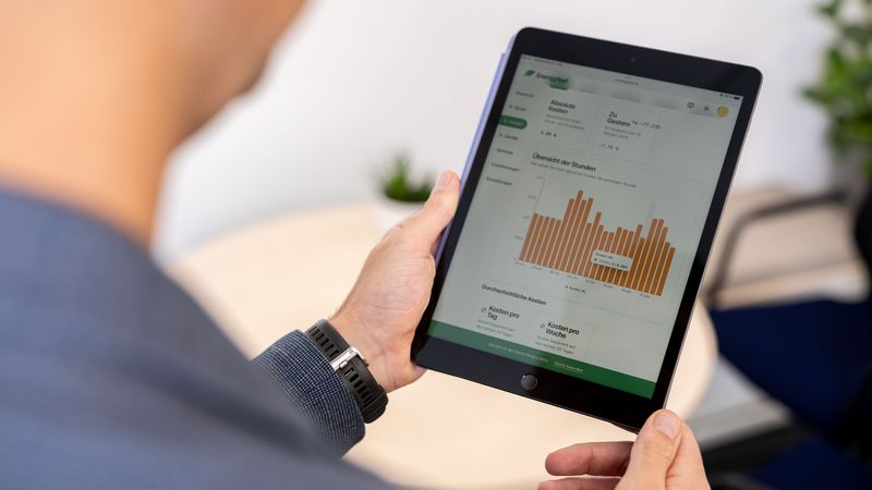 Wirtschaftsinformatiker Prof. Dr. Philipp Staudt - am Bildrand im Anschnitt unscharf abgebildet - betrachtet auf einem Tablet den Strom- und Gasbedarfs eines Beispiel-Haushalts mittels einer eigens entwickelten App. Zu sehen ist ein Balkendiagramm zum Stromverbrauch, der stundenweise aufgeschlüsselt wird, daneben verschiedene Bedienfelder, die etwa die Darstellungen der Stromkosten pro Tag oder pro Woche ermöglichen.  