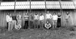 Black and white photo, a group of men, somewhat alternative-looking, standing on the lawn in front of the building, with the solar modules above them. 