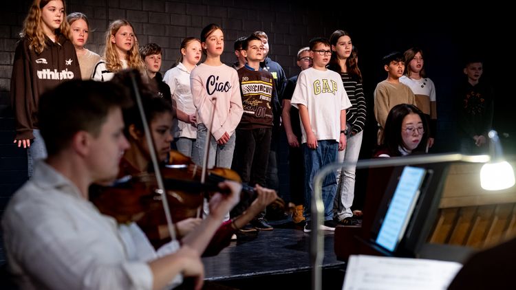 Kinder stehen auf einer Bühne und singen, davor zwei Geigenspieler.