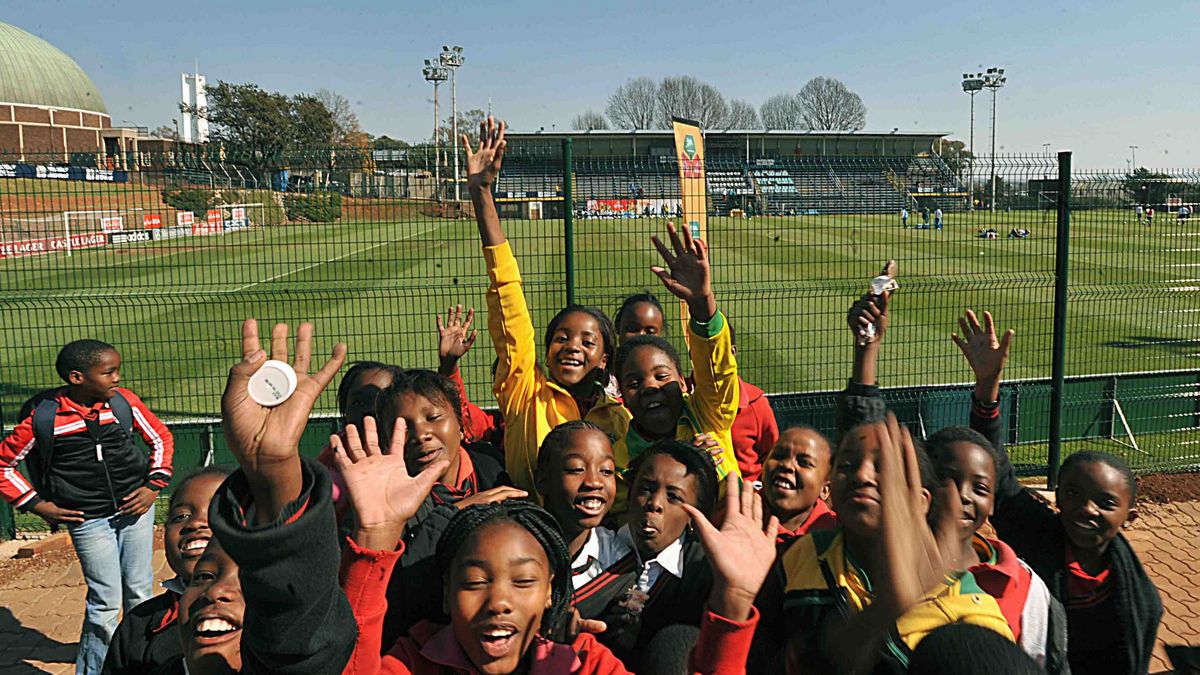 A group of children smiling and waving in front of a football stadium. A green pitch and spectator stands in the background.