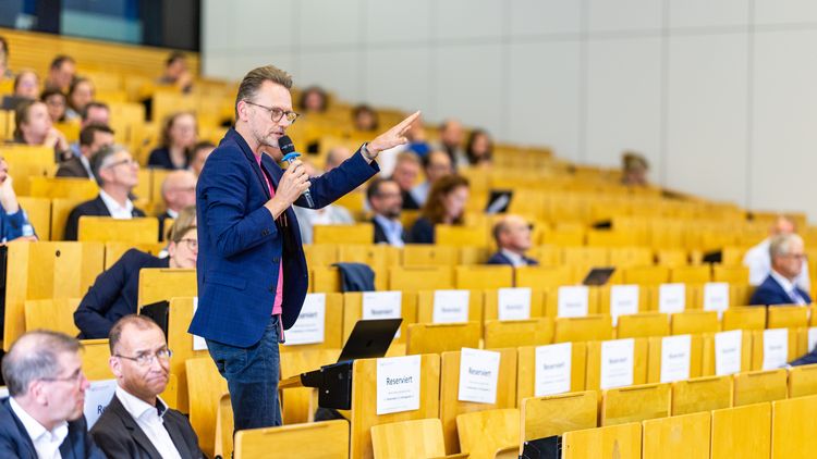A man holding a microphone is standing amongst the rows of seats in a lecture theatre, speaking and gesturing.