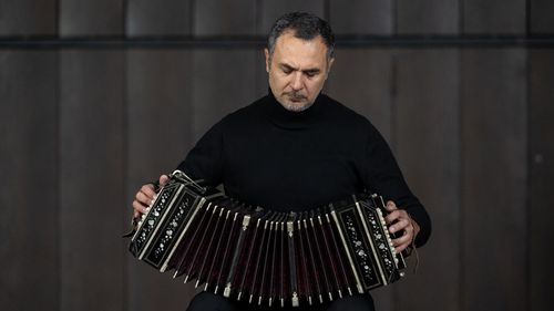 Alem sits and gazes intently down at his instrument, which he has pulled apart in front of his chest.