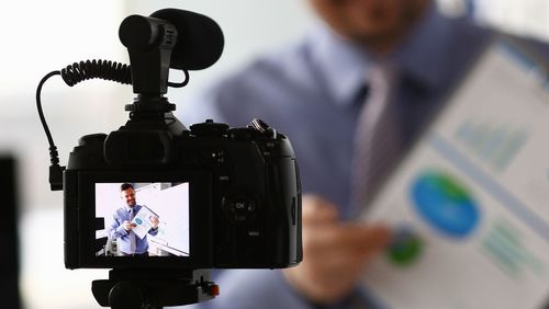 Video camera in the foreground, the display shows a man explaining a sheet of paper with some graphics.