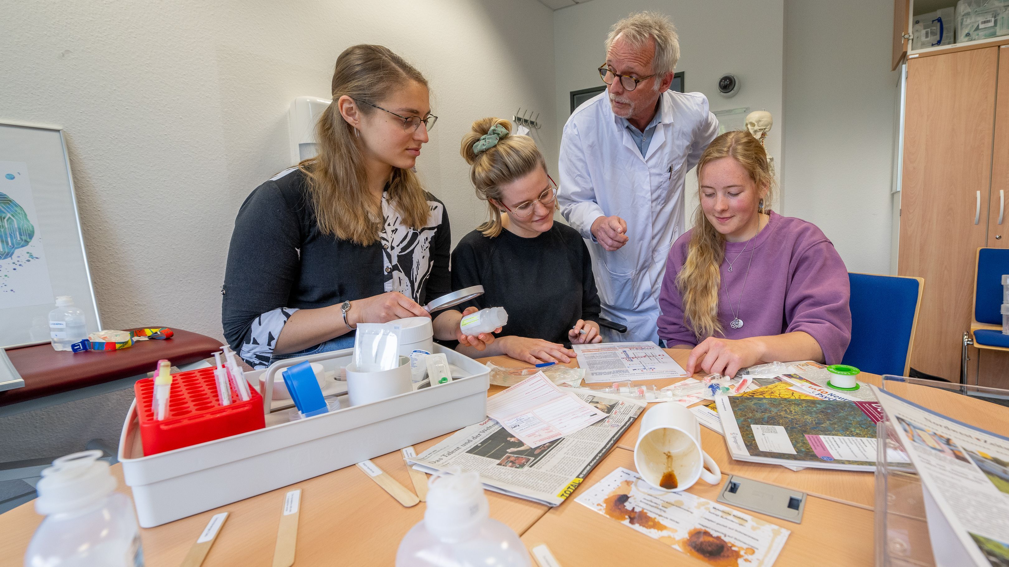 Die drei Studentinnen sitzen am Tisch, der voll ist mit medizinischen Materialien wie Mundspatel, Spritzen und Infusionsbehältern. Hinter ihnen steht ein Mann im Kittel.