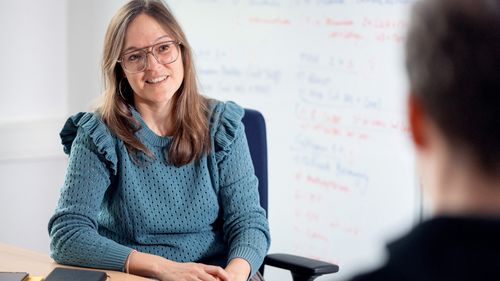 The picture shows Ineke Hess. She is sitting opposite the interviewer and looks at him. She is smiling slightly. Both are sitting at a desk. Behind Hess, a flipchart for planning can be recognised in a blur.