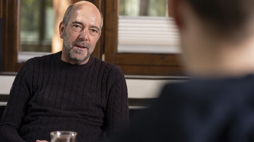 The picture shows Renke Schütte. He is sitting opposite the interviewer and looking at him. He is sitting in front of a window with a glass of tea in front of him.