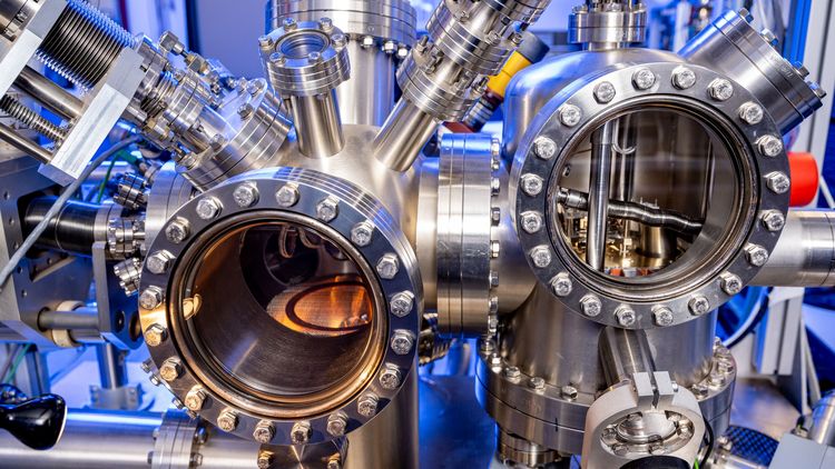 Das Bild zeigt eine physikalische Versuchsvorrichtung. Im Zentrum stehen zwei metallisches Gehäuse mit einer glatten Oberfläche, die jeweils durch eine runde Glasscheibe verschlossen sind. Sie sind etwa so groß wie ein Fußball. Die Öffnungen sehen aus wie ein Bullauge. Innerhalb der rechten Kammer befindet sich ein Wärmemikroskop, das auf dieser Totale nicht zu erkennen ist. Die Versuchsvorrichtung besteht aus einem sehr komplexen Netzwerk aus dicken Metallrohren und Kabeln.