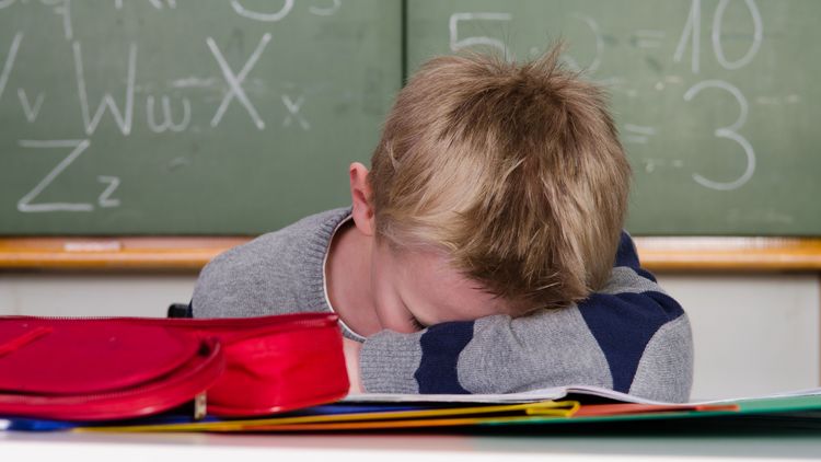 Ein Grundschüler verbirgt sein Gesicht im Arm, hinter ihm ist eine Tafel mit Zahlen und Buchstaben zu sehen.