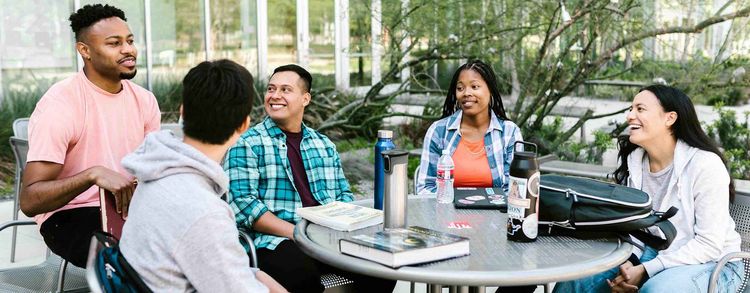 Fünf junge Menschen sitzen entspannt um einen Tisch im Freien und unterhalten sich.