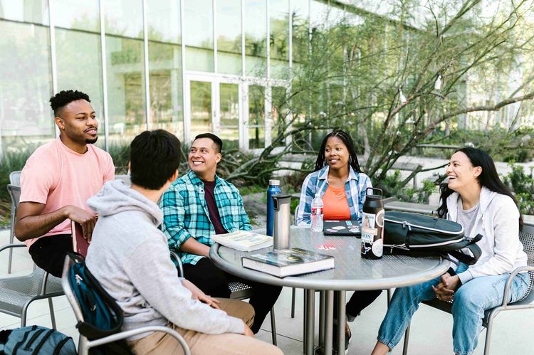 Five young people sit relaxed around a table outdoors and chat.