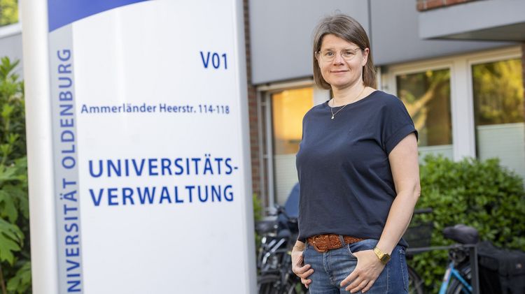 Imke Hansjürgens steht vor dem Eingang zu V01. Hinter ihr ist das große Schild zu erkennen, auf dem "Universitätsverwaltung" steht. Sie schaut in die Kamera und hat die Daumen in den Hosentaschen.