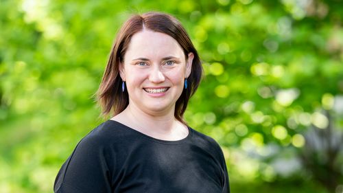Frau mit schwarzem T-Shirt und schulterlangem braunen Haar lächelt in die Kamera. Im Hintergrund sind unscharfgrüne Hecken zu sehen. 