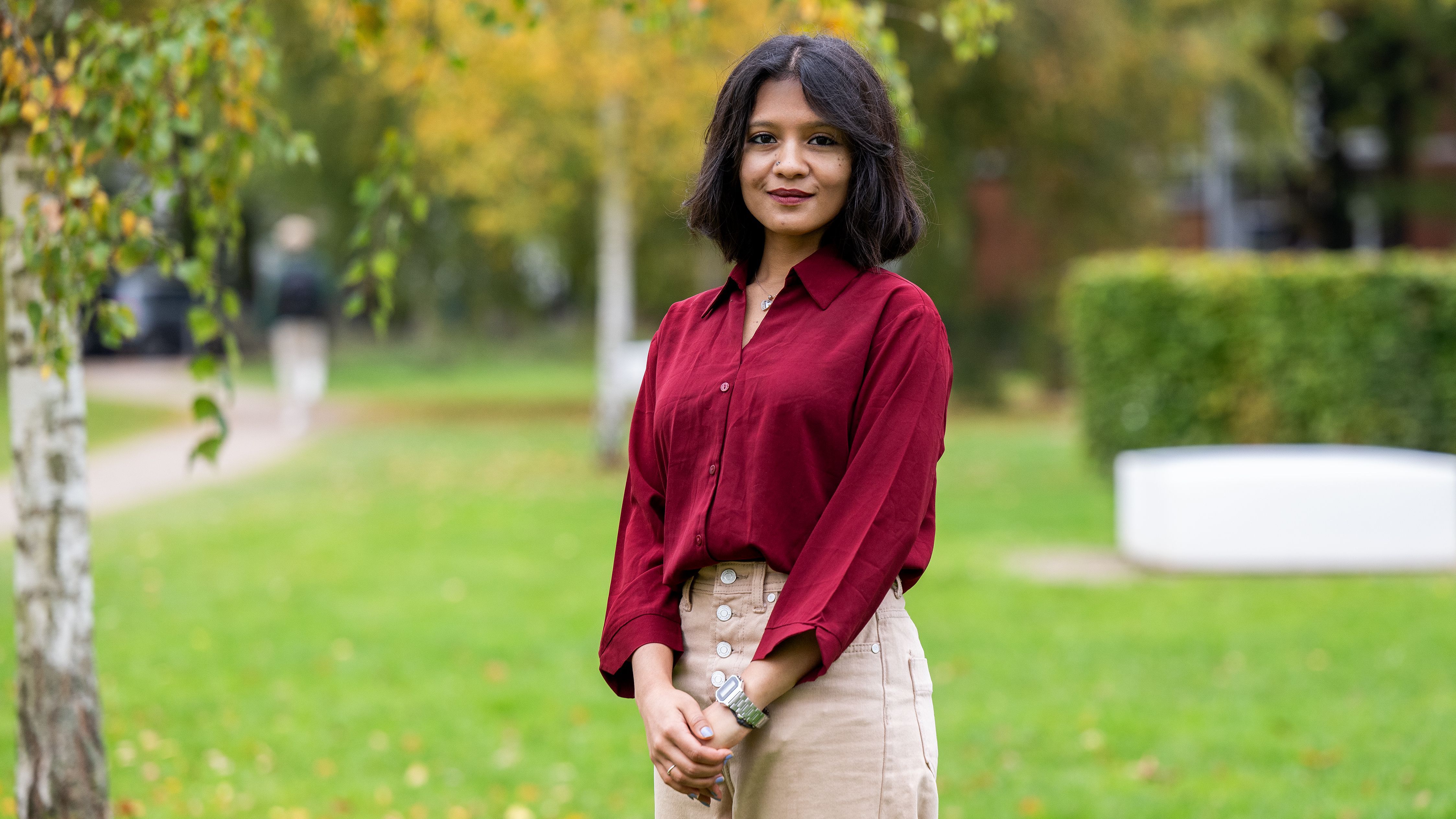 The picture shows a young adult woman wearing beige trousers and a red blouse. She is standing in a meadow, with the trunk of a birch tree visible next to her. She is smiling slightly at the camera. Her hair is shoulder-length and dark. 