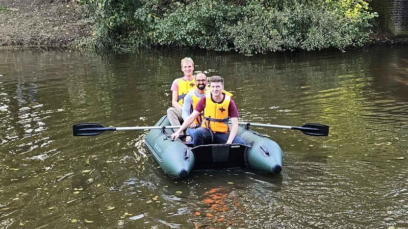 Drei Studierende des Masterstudiengangs Informatik und Wirtschaftsinformatik sitzen in einem Schlauchboot auf dem Fluss Haaren, nahe einer Brücke. Im Hintergrund sind Bäume. 