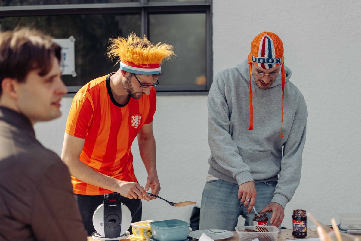 Zwei Personen mit auffälligen orangenen Mützen im Niederlande-Style bereiten Poffertjes zu.