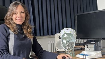 Das Bild zeigt Hanin Karawani Khoury. Sie sitzt vor einem Computer und einer Nachbildung eines menschlichen Kopfes aus Kunststoff. Ihre linke Hand hat sie auf eine Computermaus gelegt.
