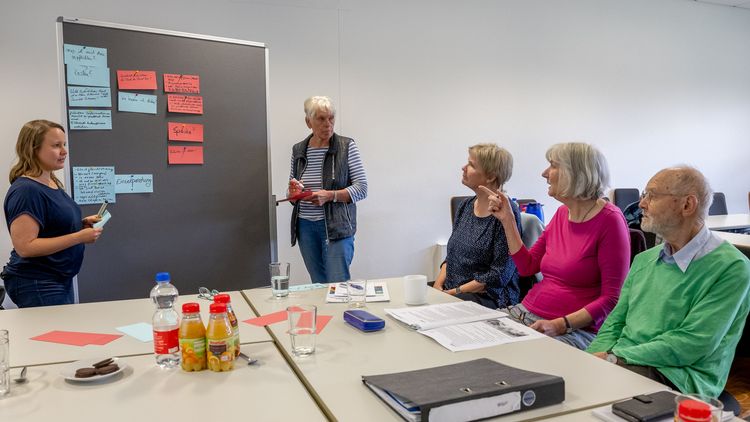 Nadja Reeck steht mit einer Seniorin an einer Pinnwannd. Beide diskutieren angeregt mit zwei älteren Damen und einem älteren Herrn, die am Tisch sitzen.
