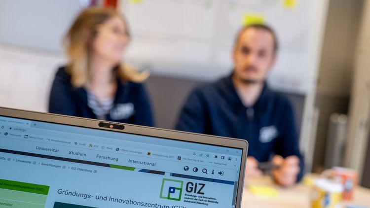 Bildschirm mit GIZ-Logo im Vordergrund, dahinter unscharf zwei Gründungsberater des GIZ.
