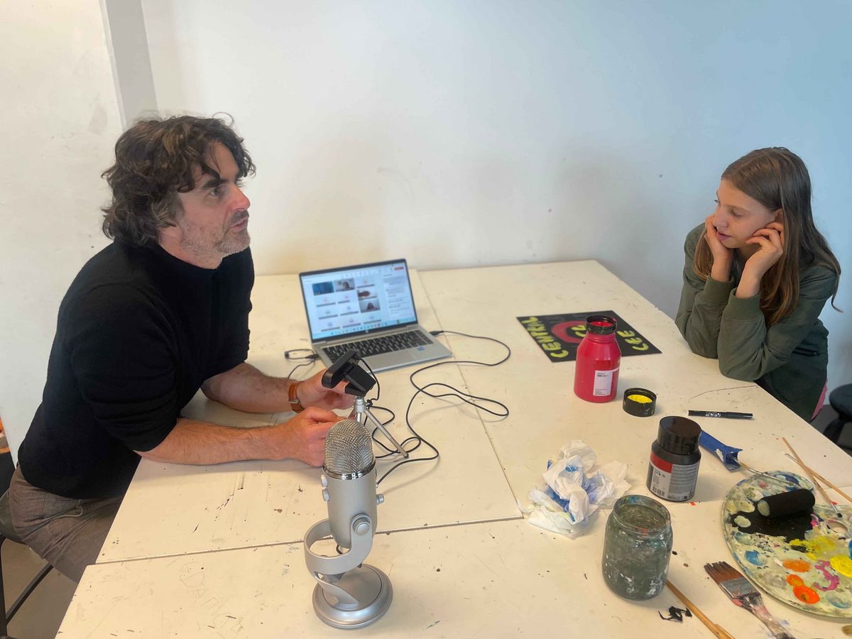 A man and a girl sit at a table and work. The man speaks into a microphone, the girl looks thoughtful.