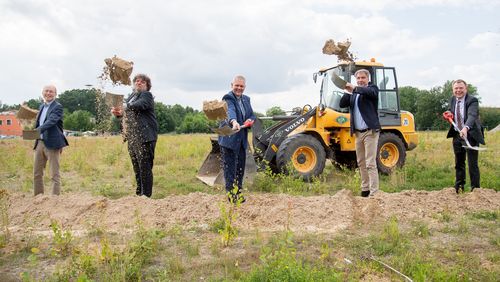 Fünf Männer mit Schaufeln stehen auf einem leeren Grundstück und werfen den geschippten Sand in die Luft. 