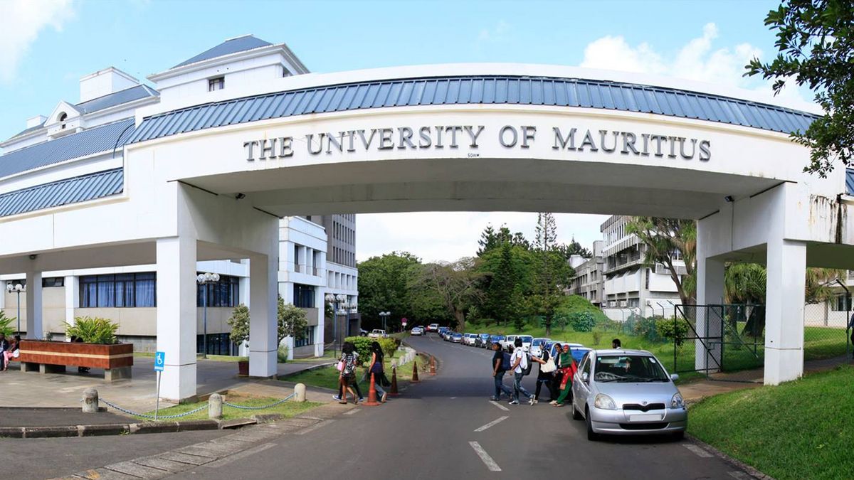 An archway with the inscription "The University of Mauritius" marks the entrance to the campus. A building with a blue and white façade can be seen behind the archway. People and vehicles can be seen on the campus.