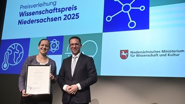 Sinikka Lennartz stands on a stage and holds a large, framed certificate in her hands. Falko Mohrs stands next to her, a screen with the Science Prize logo in the background.