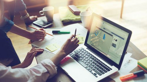 Symbolic image: Laptop on a desk, the hands of two people can be seen as well as pens, coffee cups and pieces of paper.