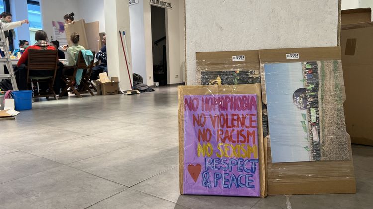 Blick in den Ausstellungsraum während des Aufbaus der Ausstellung. Im Vordergrund sind einige Bilder und Plakate zu sehen, die an einer Säule lehnen, im Hintergrund sitzen die Studierenden.