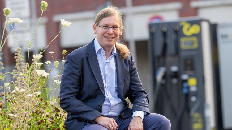 A young man with glasses and a ponytail sits on a bench and looks into the camera with a smile. Wildflowers can be seen in the background on the left and a charging point can be seen out of focus on the right.  