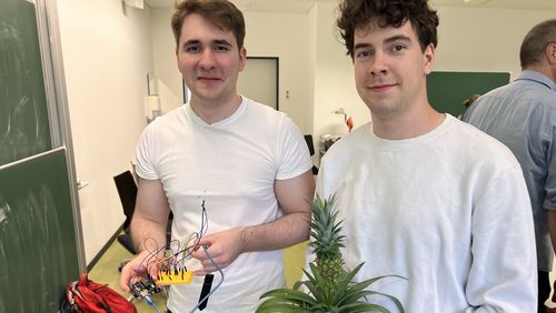Zwei Studenten blicken in die Kamera. Einer hält eine Ananaspflanze im Topf in der Hand, der andere eine gelbe Platte, aus der viele Kabel ragen.