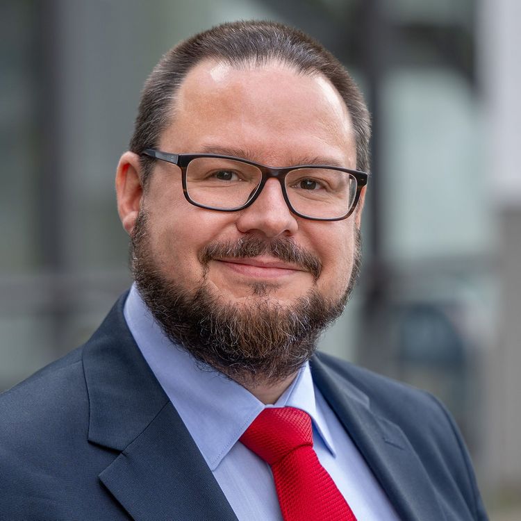 Portrait photo of Professor Dr. Torsten Jantsch, wearing black rectangular glasses, a dark blue jacket with a light blue shirt and a red tie. He smiles at the camera, has short dark hair and a short dark beard that covers his cheeks, chin and upper lip. 