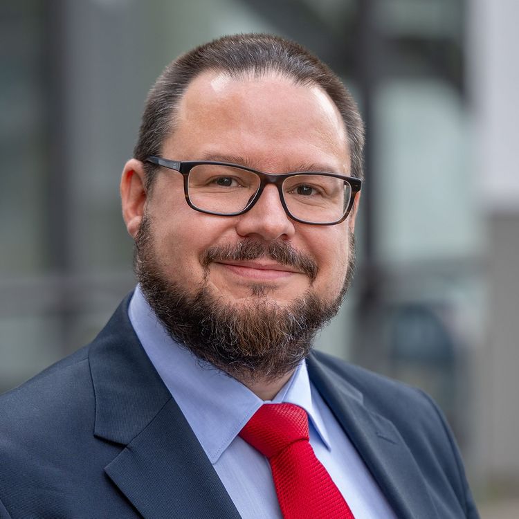 Portraitfoto von Professor Dr Torsten Jantsch, der eine schwarze rechteckige Brille, ein dunkelblaues Jacket mit hellblauem Hemd und eine rote Krawatte trägt. Er lächelt in die Kamera, hat kurze dunkle Haare und einen kurzen dunklen Bart, der Wangen, Kinn und Oberlippe bedeckt.