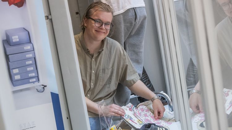 Das Bild zeigt Edwin Mårtensson. Er sitzt auf einer Auslage hinter einem Schaufenster und ist von diversen Textilien, Schuhen und anderen Gegenständen umgeben. Er lächelt in die Kamera.