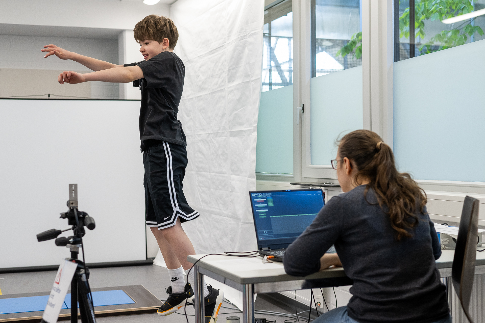 Child jumps into the air. Adult sits behind laptop and watches boy.