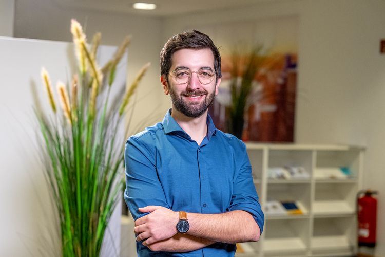 Das Bild zeigt Can Eroglu. Er steht mit verschränkten Armen vor einem Wandbild, das die Uni Oldenburg im Winter zeigt. Er trägt ein blaues Hemd, Brille und lächelt in die Kamera.