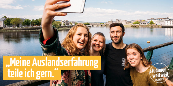 Ein Selfie von vier jungen Menschen vor einer Stadtlandschaft mit einem Fluss und Brücken. Die Personen lächeln in die Kamera. Auf dem Bild befindet sich ein gelber Balken mit dem Text: „Meine Auslandserfahrung teile ich gern.“ Rechts unten ist ein Logo mit dem Slogan „studieren weltweit – erlebe es!“ zu sehen. 