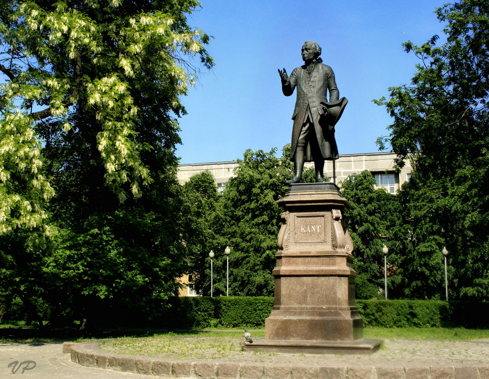 Das Bild zeigt ein Denkmal für Immanuel Kant. Kant steht auf einem Sockel und blickt in die Ferne. Im Hintergrund sind ein Park und Gebäude zu erkennen.