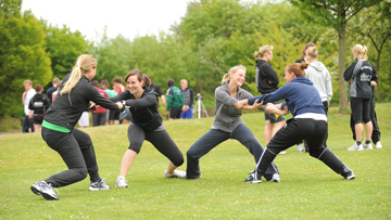 Das Bild zeigt eine Gruppe Studierende auf einer Grünfläche, die sich in Zweiergruppen gegenseitig an den Armen halten und einer sportlichen Aktivität nachgehen.