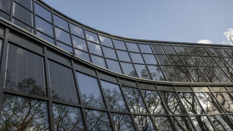 Das Foto zeigt einen Ausschnitt der modernisierten Glasfassade. Man kann in einigen der einzelnen Glasscheiben die integrierten PV-Module erkennen. Außerdem spiegeln sich auf dem Glas die umstehenden Bäume.